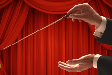 The hands of a conductor with a baton in front of a red-curtained stage.