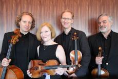 The American String Quartet dressed in concert black standing in front of a wooden background with their instruments, 
