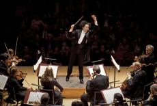 Los Angeles Philharmonic orchestra conductor with his arms raised in front of orchestra. 