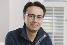 Conductor Ken David-Masur posing indoors seated, wearing a blue sweater