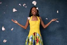 Isata poses in a yellow sleeveless dress with her arms open in front of a dark blue background, pink flower petals are falling.