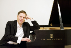 Alon Goldstein sits at a grand piano in a black suite and white shirt, resting his head on his left hand. 