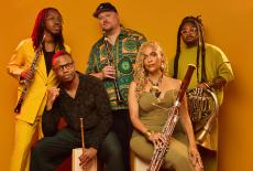 The wind quintet Imani Winds poses in front of a burnt orange background with their instruments.