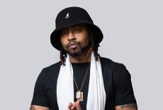 Wordsmith poses in front of a white background, he is wearing black, a white scarf, and black bucket hat