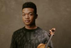 Randall poses with his violin in his left arm against his body, wearing a darkly colored animal print sweater.