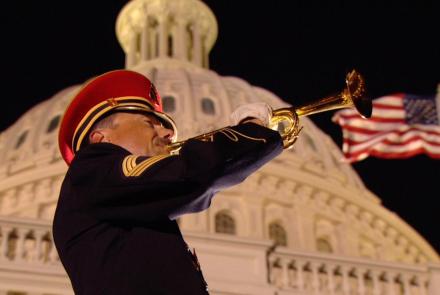 A soldier plays a trumpet in front of the US Capitol with the American flag flying.