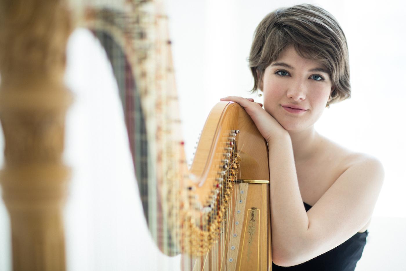 Harpist Emily Levin posing with her head on her hand resting on her harp in front of a white background.