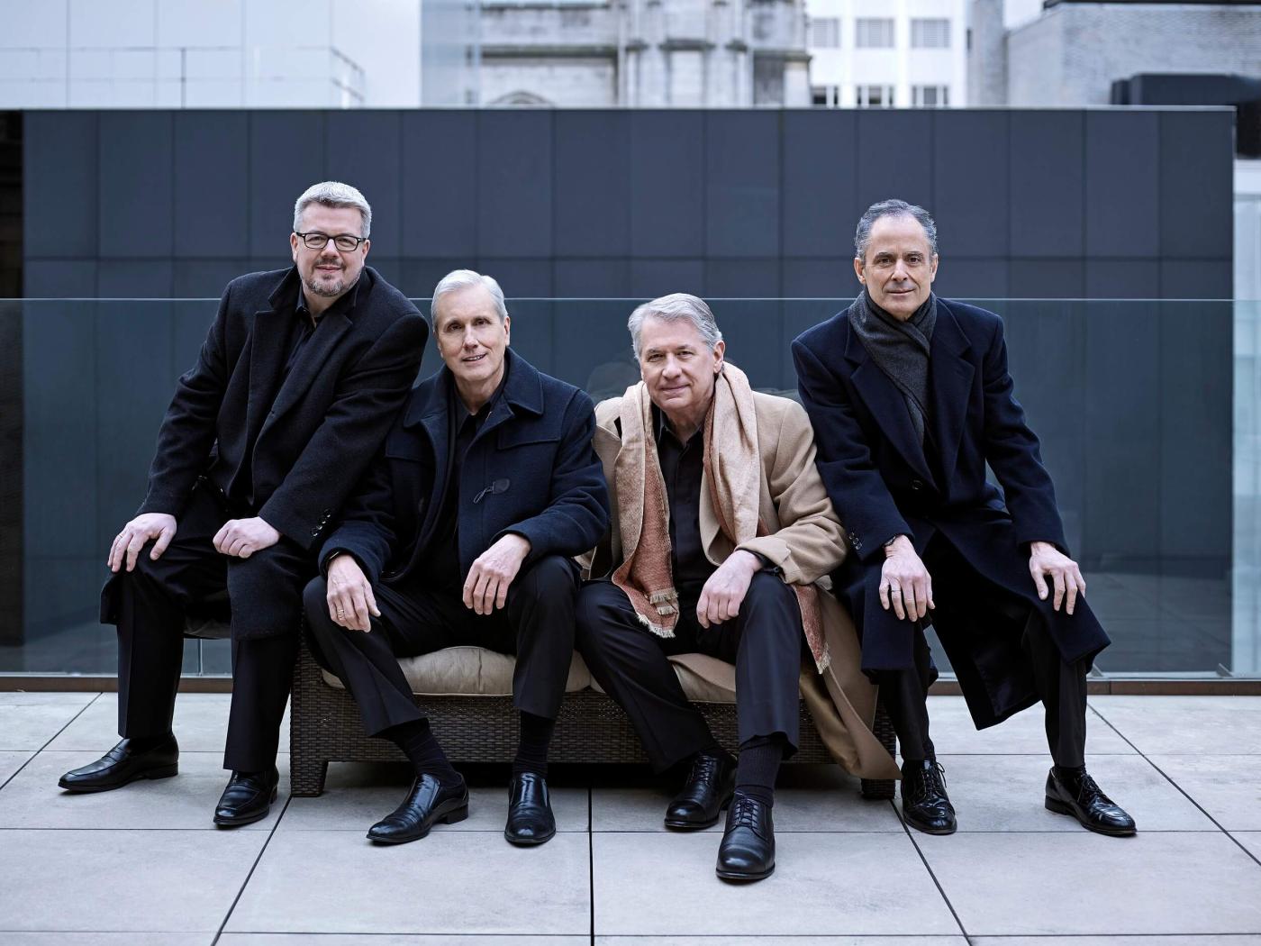 The Emerson Quartet poses outside on the sidewalk, sitting on a concrete bench dressed in black. 