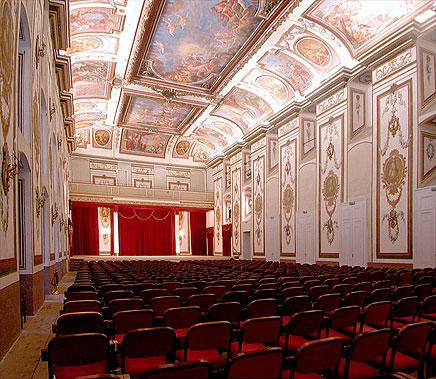 A view of Haydn's concert hall at Esterhazy. The walls are white with gold leaf, frescos on the ceiling, and red chairs for the audience.